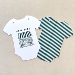May include: Two baby bodysuit-shaped paper cutouts. One is white with a black and white cafe illustration and the text "CAFE BEBE". The other is green and white striped.