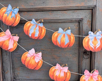 May include: A festive pumpkin garland with orange pumpkins and blue or pink bows. The garland is strung on a white string and is perfect for fall or Thanksgiving decorations. The pumpkins have green stems.