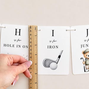 May include: A white rectangular card featuring the letter H, the text 'is for HOLE IN ONE', and a wooden ruler measuring approximately 20 cm in height. Next to it is a card with the letter I, the text 'is for IRON', and a golf club illustration.