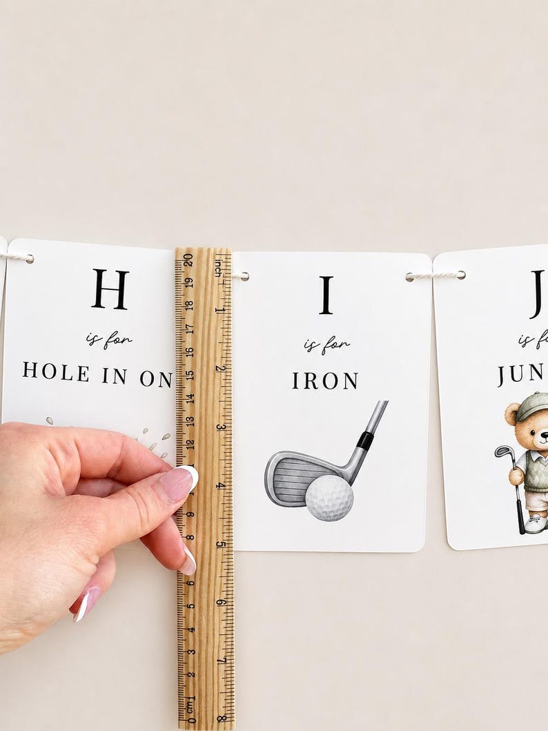 May include: A white rectangular card featuring the letter H, the text 'is for HOLE IN ONE', and a wooden ruler measuring approximately 20 cm in height. Next to it is a card with the letter I, the text 'is for IRON', and a golf club illustration.