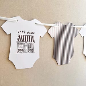 May include: White paper cutouts in the shape of baby onesies hang from a white string. One features a black and white illustration of a cafe with the text "CAFE BEBE". Another has black and white vertical stripes.