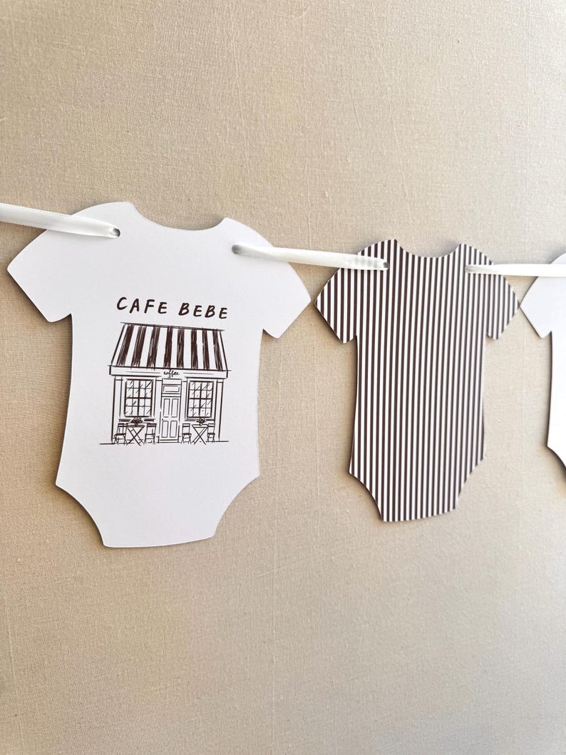 May include: White paper cutouts in the shape of baby onesies hang from a white string. One features a black and white illustration of a cafe with the text "CAFE BEBE". Another has black and white vertical stripes.