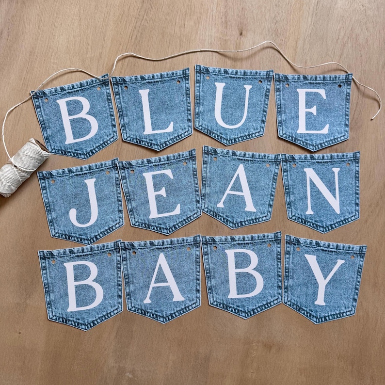 May include: A decorative banner consisting of twelve individual denim pocket-shaped fabric pieces strung together on a white cord. Each pocket features a large white capital letter, collectively spelling out the phrase BLUE JEAN BABY.