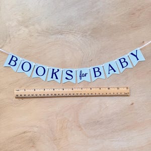 K&ouml;nnte beinhalten: Ein Banner mit der Aufschrift "BOOKS for BABY" in marineblauen Buchstaben auf hellblau-karierten F&auml;hnchen. Unter dem Banner befindet sich ein Holzlineal. Das Banner ist an einer wei&szlig;en Schnur befestigt.