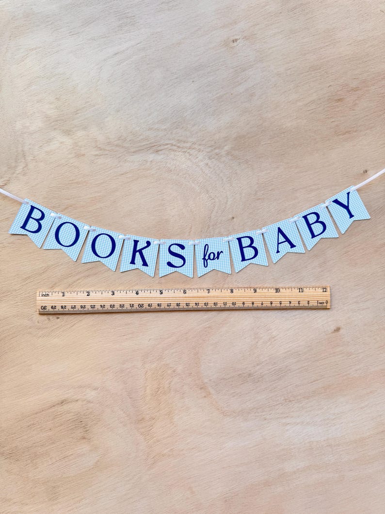 K&ouml;nnte beinhalten: Ein Banner mit der Aufschrift "BOOKS for BABY" in marineblauen Buchstaben auf hellblau-karierten F&auml;hnchen. Unter dem Banner befindet sich ein Holzlineal. Das Banner ist an einer wei&szlig;en Schnur befestigt.
