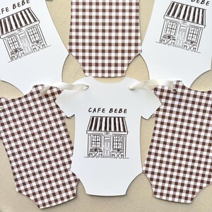 May include: Baby bodysuit-shaped paper cutouts. Some are white with a black line drawing of a cafe and the words "CAFE BEBE". Others are brown and white checkered. White ribbon is attached to some of the cutouts.