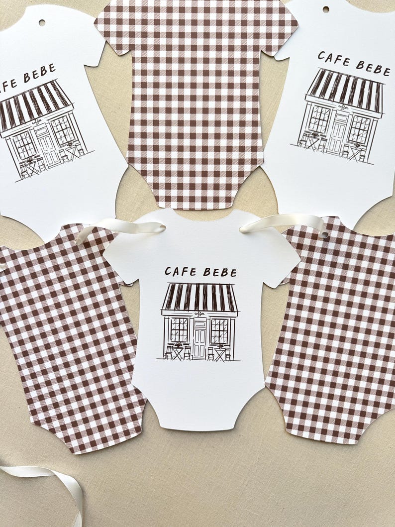 May include: Baby bodysuit-shaped paper cutouts. Some are white with a black line drawing of a cafe and the words "CAFE BEBE". Others are brown and white checkered. White ribbon is attached to some of the cutouts.