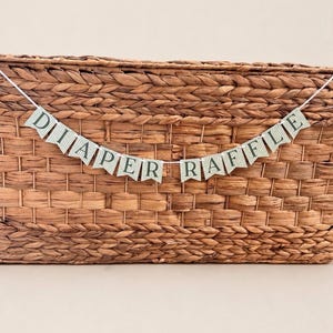 K&ouml;nnte beinhalten: Ein Banner mit der Aufschrift "DIAPER RAFFLE" auf kleinen, gr&uuml;n-wei&szlig; karierten F&auml;hnchen. Es ist &uuml;ber einem geflochtenen Korb gespannt und eignet sich als festliche Dekoration f&uuml;r eine Babyparty oder &auml;hnliche Veranstaltung.