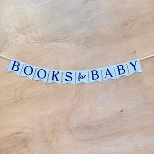 K&ouml;nnte beinhalten: Ein Banner mit der Aufschrift "BOOKS for BABY" in Marineblau auf hellblauen Wimpeln mit Vichy-Muster. Der Banner ist auf einer wei&szlig;en Schnur aufgereiht und vor einem hellbraunen Holzgrund platziert.