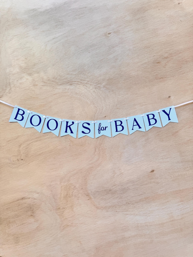 K&ouml;nnte beinhalten: Ein Banner mit der Aufschrift "BOOKS for BABY" in Marineblau auf hellblauen Wimpeln mit Vichy-Muster. Der Banner ist auf einer wei&szlig;en Schnur aufgereiht und vor einem hellbraunen Holzgrund platziert.