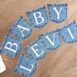 May include: A decorative banner composed of eight individual blue denim pockets arranged in two rows. Each pocket features a single large white capital letter, collectively spelling out the words BABY LEVI. Each pocket has two small holes for stringing.