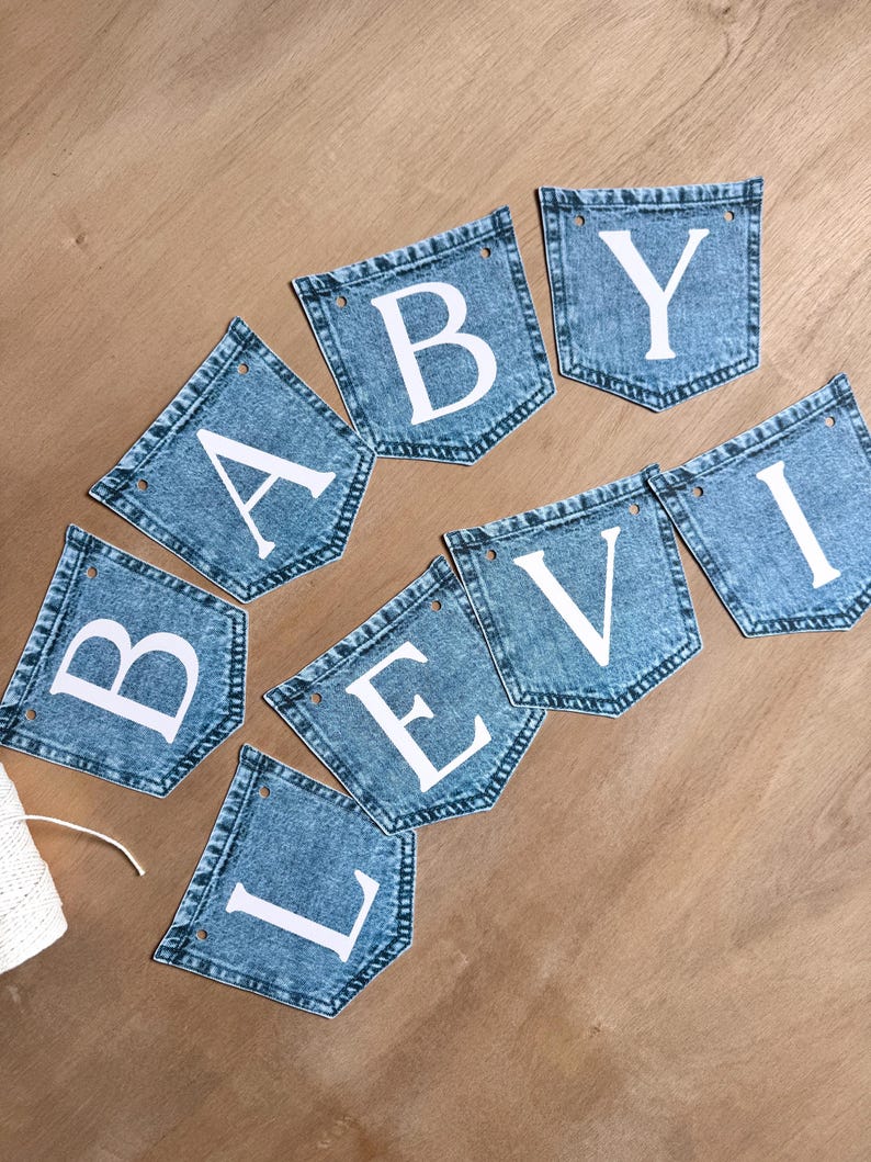 May include: A decorative banner composed of eight individual blue denim pockets arranged in two rows. Each pocket features a single large white capital letter, collectively spelling out the words BABY LEVI. Each pocket has two small holes for stringing.