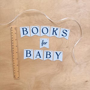 K&ouml;nnte beinhalten: Ein Banner mit der Aufschrift "BOOKS for BABY" in dunkelblauen Buchstaben auf hellblau-wei&szlig; kariertem Hintergrund. Ein Holzlineal liegt neben dem Banner, und ein wei&szlig;es Band ist &uuml;ber die Holzoberfl&auml;che drapiert.