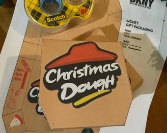 Puede incluir: Una caja de pizza de cartón marrón con las palabras "Christmas Dough" en blanco y negro, con un logotipo rojo y amarillo. Un rollo de cinta adhesiva Scotch está encima de la caja y una hoja de papel con instrucciones es visible.