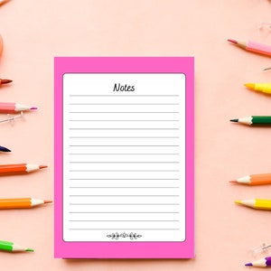 Op de afbeelding: Een roze notitieboekje met gestreept papier en het woord "Notes" bovenaan. Het notitieboekje is omringd door kleurpotloden en duimspijkers op een roze achtergrond.