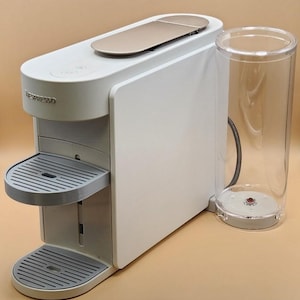 May include: A white Nespresso coffee machine with a gold-colored top and a clear water reservoir. The machine has two gray drip trays and the Nespresso logo on the front. The machine is designed for making coffee.