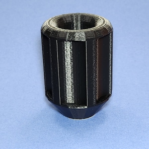 May include: A black 3D printed object with a cylindrical shape and a pointed bottom. The object has a series of vertical lines around its circumference.