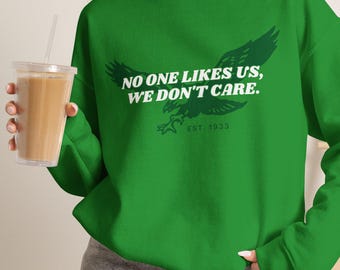 Może przedstawiać: Jaskrawozielona bluza z długim rękawem i okrągłym dekoltem, ozdobiona ciemnozieloną grafiką orła oraz białym napisem "NO ONE LIKES US, WE DON'T CARE." i "EST. 1933" na klatce piersiowej. Bluza posiada ściągacze przy kołnierzu, mankietach i u dołu.