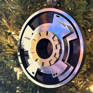 May include: A round, layered Christmas ornament with a metallic silver, black, and gold design. The ornament has a complex, geometric pattern and is hanging from a silver ribbon against a backdrop of green Christmas tree branches.