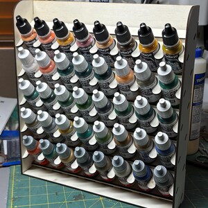 May include: A wooden paint bottle organizer filled with various colors of Vallejo Model Color paints. The organizer has multiple angled shelves to hold the bottles. The paint colors include shades of red, yellow, green, blue, and brown.