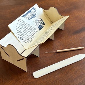 May include: A light-colored wooden book cradle with a curved side profile holding an open booklet with black ink illustrations and handwritten text. Beside the cradle lies a thin wooden-handled metal awl and a smooth white plastic bone folder.