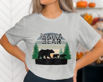 Puede incluir: Una camiseta gris con un gráfico de una osa madre y dos cachorros en un entorno de bosque. El texto "Mama Bear" está impreso en negro sobre el gráfico.