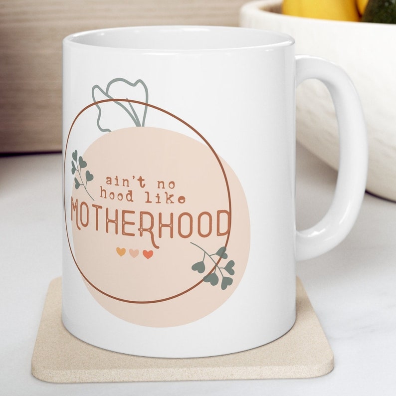 May include: White ceramic mug with the text "ain't no hood like MOTHERHOOD" in a brown, peach, and green design. The mug has a curved handle and sits on a beige coaster. The design includes a circle, hearts, and floral elements.