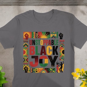 Può includere: T-shirt grigia con la scritta "UNSTOPPABLE BLACK JOY" in un design colorato. Il design include un pugno alzato, raggi di sole e motivi geometrici in rosso, verde, giallo e nero. Sono incluse anche due figure danzanti.