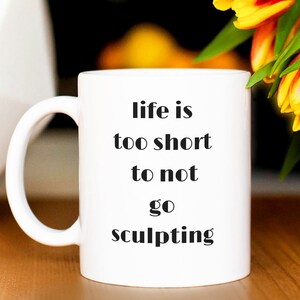 May include: White ceramic mug with a black inscription that reads "life is too short to not go sculpting". The mug is sitting on a wooden table next to a bouquet of orange and yellow tulips.