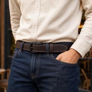 May include: A dark brown leather belt with visible stitching along the edges and a matte black metal buckle. The belt is worn through the belt loops of dark blue denim jeans, paired with a long-sleeved, button-down cream-coloured shirt.
