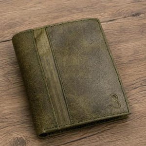 May include: A bi-fold wallet in a distressed olive green finish with a vertical textured panel on the left side. The wallet features a smooth, matte surface and a small, embossed logo of a stylized ram head in the bottom right corner.