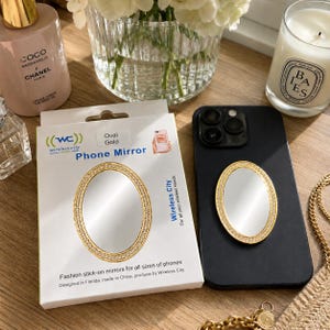 May include: An oval gold phone mirror with a rhinestone-encrusted border, packaged in a white box labeled Wireless City Phone Mirror, alongside a black smartphone with an identical mirror attached to its back surface for mobile device use.