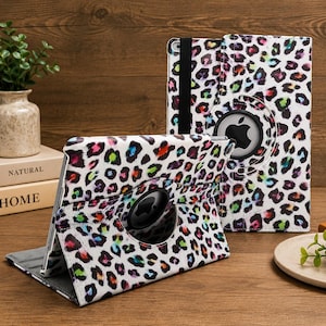 May include: A white tablet case featuring a multicolored leopard print pattern with black, pink, blue, and green spots. The case includes a circular cutout to display the Apple logo and a black elastic strap for secure closure when folded.
