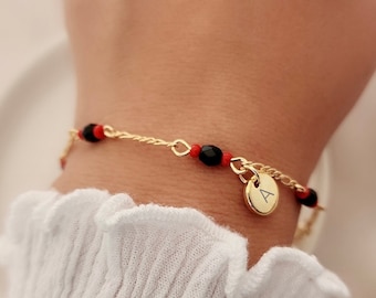 May include: A gold chain bracelet with black and red beads and a gold charm with the letter 'A' on it.