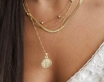 May include: A gold layered necklace with a round pendant featuring a detailed design. The necklace is worn over a white lace top.
