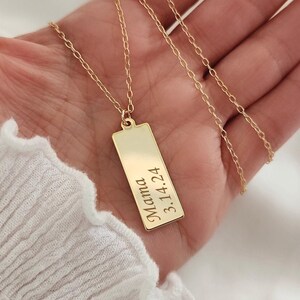 May include: A gold-colored rectangular pendant necklace with the word "Mama" and the date "3.14.24" engraved on it. The necklace is held in a hand, showcasing the delicate chain and pendant design. The background is a soft, white fabric.