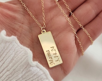 May include: A gold-colored rectangular pendant necklace with the word "Mama" and the date "3.14.24" engraved on it. The necklace is held in a hand, showcasing the delicate chain and pendant design. The background is a soft, white fabric.
