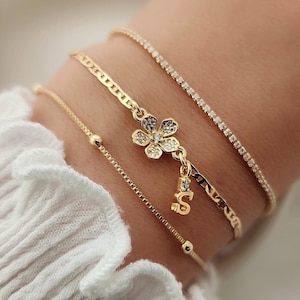 May include: Four gold bracelets stacked on a wrist. The top bracelet is a thin, sparkly chain. The second bracelet is a thicker chain with a small flower charm. The third bracelet is a thin chain with a small bead at the end. The bottom bracelet is a thin, sparkly chain.
