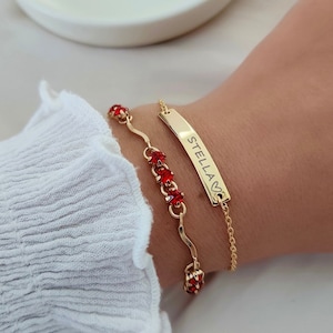 Puede incluir: Dos pulseras de oro en una muñeca. Una pulsera tiene una cadena fina con piedras preciosas rojas y la otra tiene una cadena más gruesa con una barra de oro que dice "STELLA".