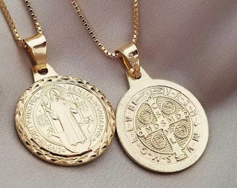 May include: Two gold-colored Saint Benedict medallions on delicate chains. The pendants feature detailed engravings of religious figures and symbols. The chains and pendants are displayed on a neutral background.