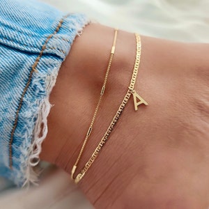 May include: Two delicate gold anklets. One is a simple chain, and the other features a small letter "A" pendant. The anklets are worn on an ankle, with a light blue denim jean visible in the background.