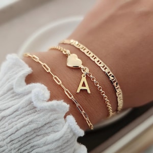 May include: A gold chain bracelet with a heart charm and a letter 'A' charm. The bracelet is layered with two other gold chain bracelets.