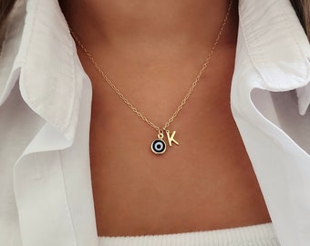 May include: Gold-colored necklace with a delicate chain. The necklace features a small, round charm with a blue and white eye design, and a gold letter "K" charm. The necklace is worn against a white shirt.