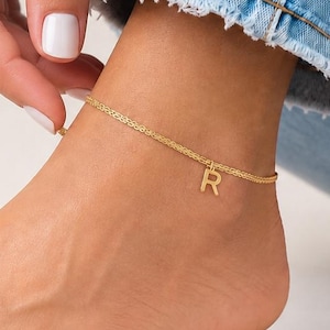 Puede incluir: Una tobillera dorada con un dije de la letra "R". La delicada cadena se lleva en un tobillo de piel clara, combinada con unos vaqueros azules. La tobillera es una joya sencilla y elegante.
