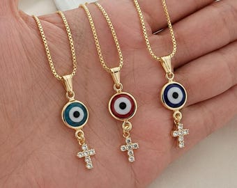 May include: Three gold-colored necklaces with pendant charms. Each necklace features a round evil eye charm in blue, red, and blue, and a small cross pendant with clear crystals. The necklaces are displayed on a light-colored surface.