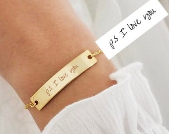 Puede incluir: Pulsera de cadena dorada con una etiqueta rectangular grabada con el mensaje escrito a mano "p.s. Te quiero".