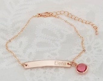 May include: Rose gold bracelet with a bar engraved with the name "Elena" and a dangling round pink gemstone. The bracelet is on a white lace surface with a pink ribbon in the background.