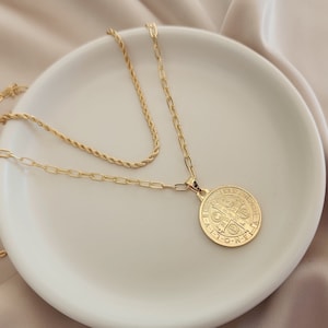 May include: A gold chain necklace with a round pendant featuring a detailed design. The necklace is layered on a white plate.