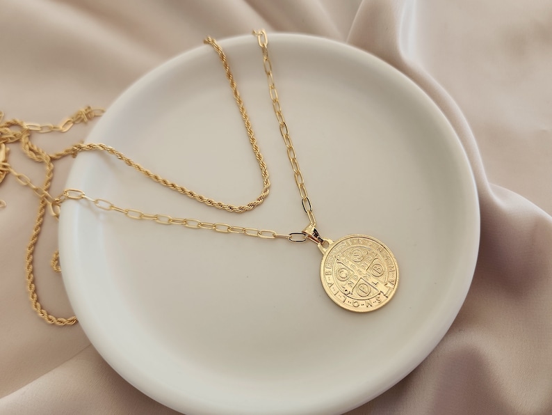 May include: A gold chain necklace with a round pendant featuring a detailed design. The necklace is layered on a white plate.