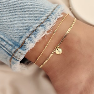 Op de afbeelding: Gouden enkelband met een klein bedeltje met de letter 'A' erop. De enkelband is gelaagd met een dunnere ketting.